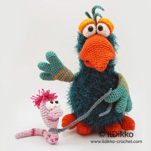 May include: A teal fuzzy crocheted bird with a large orange beak, orange feet, and wide eyes, holding a gray leash attached to a small pink crocheted worm with pink hair. The text "© IlDikko www.ildikko-crochet.com" is printed at the bottom.