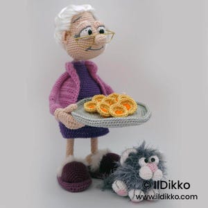 May include: A handmade crochet doll of an elderly woman wearing a purple dress and pink jacket, holding a plate of crocheted pastries. A gray cat with pink accents sits at her feet. The doll has glasses and white hair. The image is set against a white background.