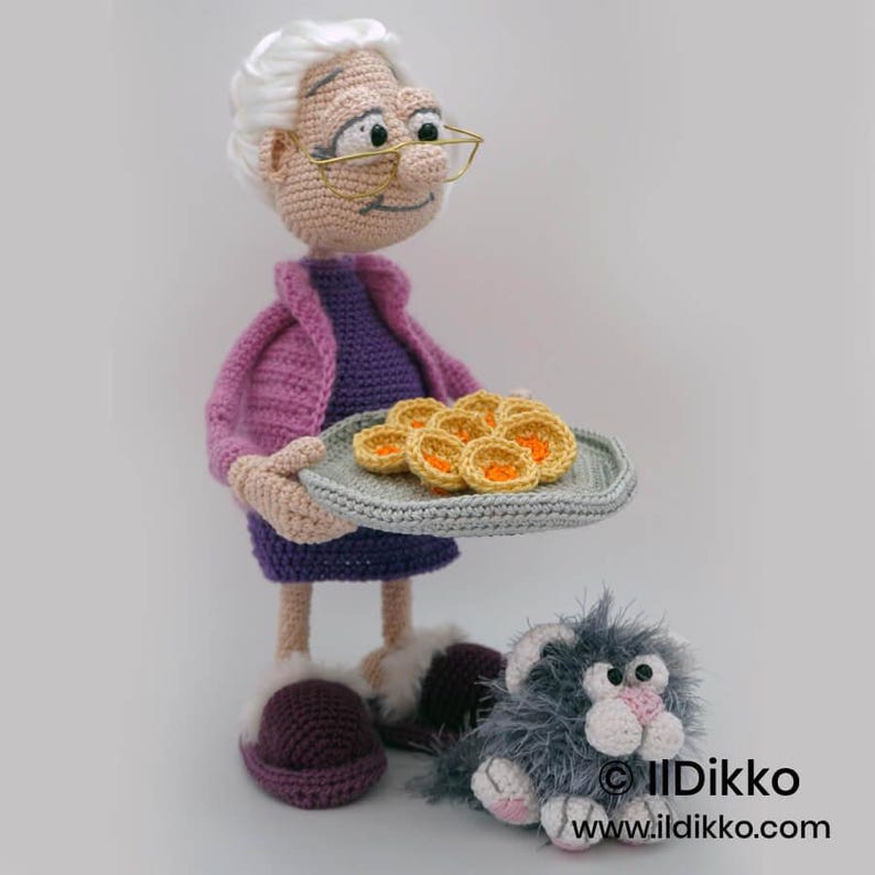 May include: A handmade crochet doll of an elderly woman wearing a purple dress and pink jacket, holding a plate of crocheted pastries. A gray cat with pink accents sits at her feet. The doll has glasses and white hair. The image is set against a white background.