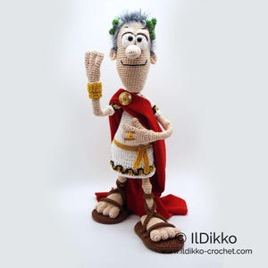 以下が含まれることがあります： 白いトガ、赤いマント、金のジュエリーを身に着けたローマ皇帝の、かぎ針編みアミグルミ人形。人形は灰色のひげと頭の上に月桂樹の輪を持っています。人形は右手を上げて挨拶をしています。
