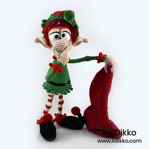 May include: A crocheted elf doll with red hair, a green hat with sequins, and a green tunic. The doll has striped red and white legs, black curled shoes with red pom-poms, and holds a red textured stocking. Text reads: &copy; Ildikko www.ildikko.com