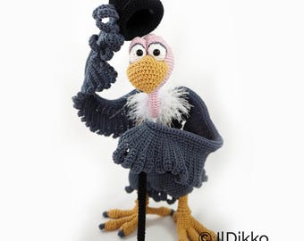 May include: A crocheted vulture figurine with a pink head, yellow beak, and white neck feathers. The vulture has dark gray wings, yellow feet with dark claws, and holds a black top hat in one wing while leaning on a thin black cane. Text reads IlDikko.
