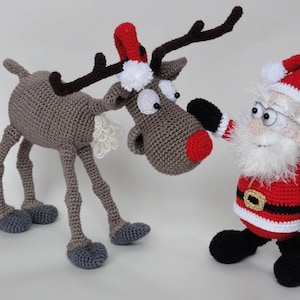 Puede incluir: Un reno de ganchillo con nariz roja, cuerpo marrón y astas oscuras que lleva un pequeño gorro rojo, junto a una figura de Papá Noel de ganchillo con barba blanca, traje rojo, botas negras y un gorro rojo con un pompón blanco.