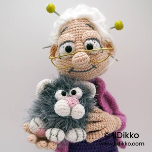 May include: A crocheted doll of a smiling elderly woman with white hair, glasses, and knitting needles. She is holding a gray and white crocheted cat with a pink nose. The doll is wearing a purple outfit and a pink sweater.