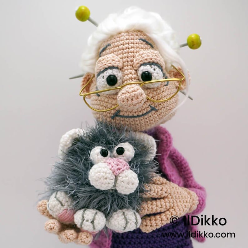 May include: A crocheted doll of a smiling elderly woman with white hair, glasses, and knitting needles. She is holding a gray and white crocheted cat with a pink nose. The doll is wearing a purple outfit and a pink sweater.