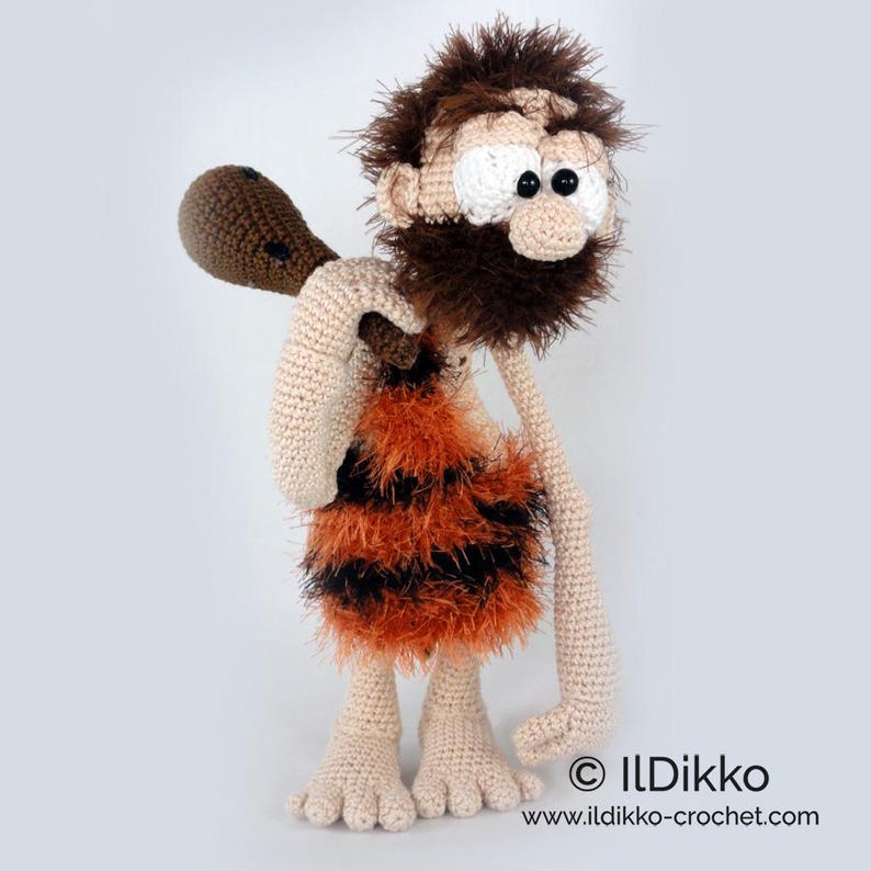 May include: A crocheted caveman doll with beige skin, a shaggy brown beard, and large white eyes with black pupils. The figure wears a fuzzy orange and black striped tunic and carries a brown club over one shoulder. Text reads ILDikko www.ildikko-crochet.com.