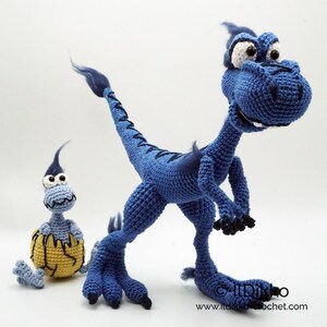 May include: A large blue crocheted dinosaur with dark blue accents and a smaller light blue crocheted dinosaur emerging from a yellow cracked egg. The text "IlDikko" and "www.itdikko.crochet.com" is printed in the bottom right corner.