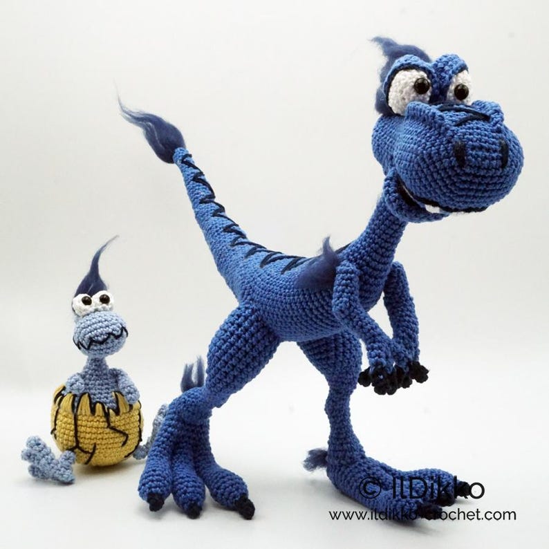 May include: A large blue crocheted dinosaur with dark blue accents and a smaller light blue crocheted dinosaur emerging from a yellow cracked egg. The text "IlDikko" and "www.itdikko.crochet.com" is printed in the bottom right corner.
