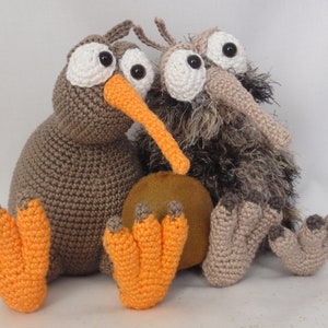 Puede incluir: Dos peluches de kiwi de crochet marrón con patas naranjas y grandes ojos blancos. El kiwi de la derecha tiene pelo marrón y está sentado sobre un huevo de fieltro marrón.