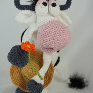 May include: A crocheted cow toy with black and white spots, pink nose, and black eyes. The cow is holding a brown hat and a green stem with an orange flower.