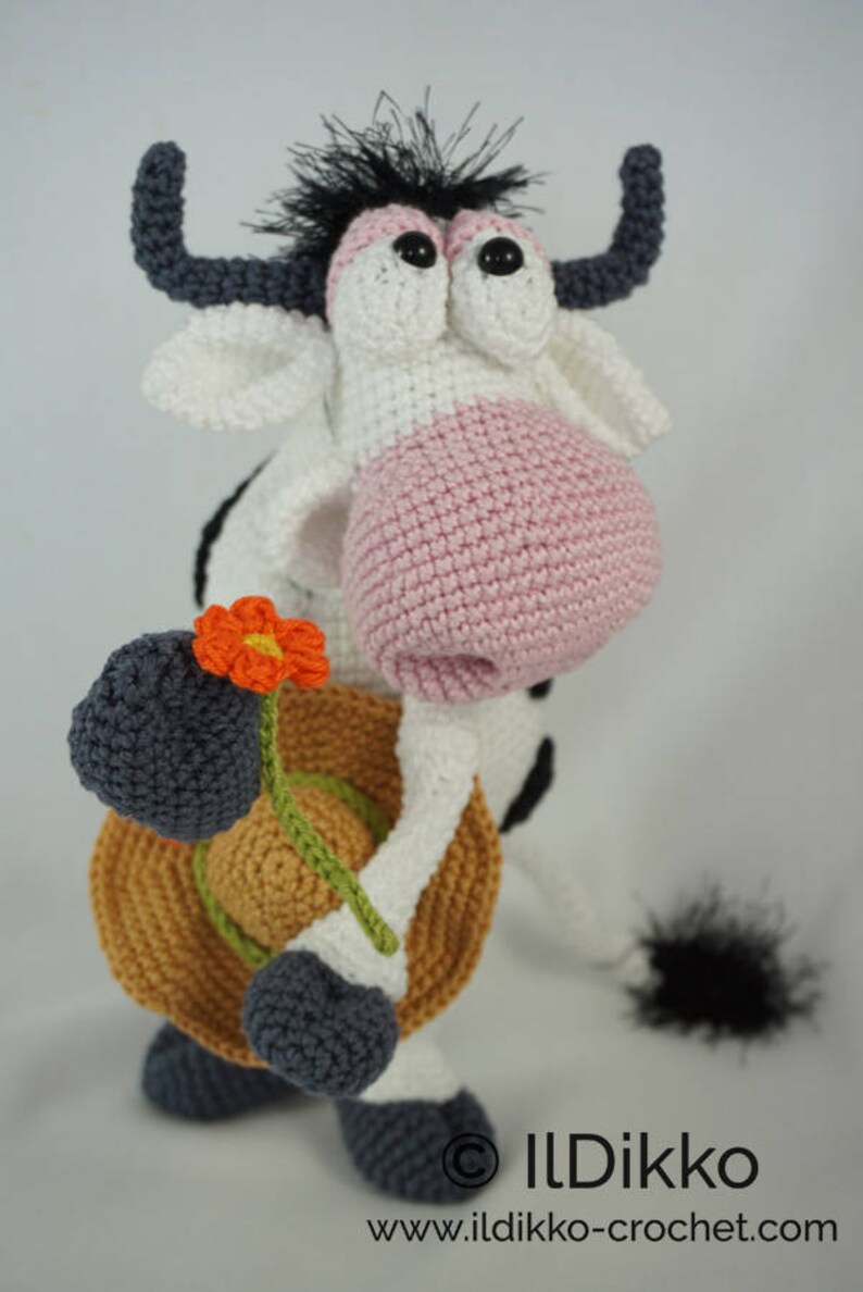 May include: A crocheted cow toy with black and white spots, pink nose, and black eyes. The cow is holding a brown hat and a green stem with an orange flower.