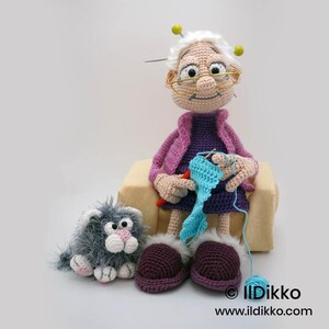 May include: A crocheted doll of an elderly woman wearing glasses, a purple dress, and a pink jacket, crocheting with blue yarn. A gray cat and purple slippers with white fur trim are nearby. The doll has white hair and yellow pins. The website address is visible.