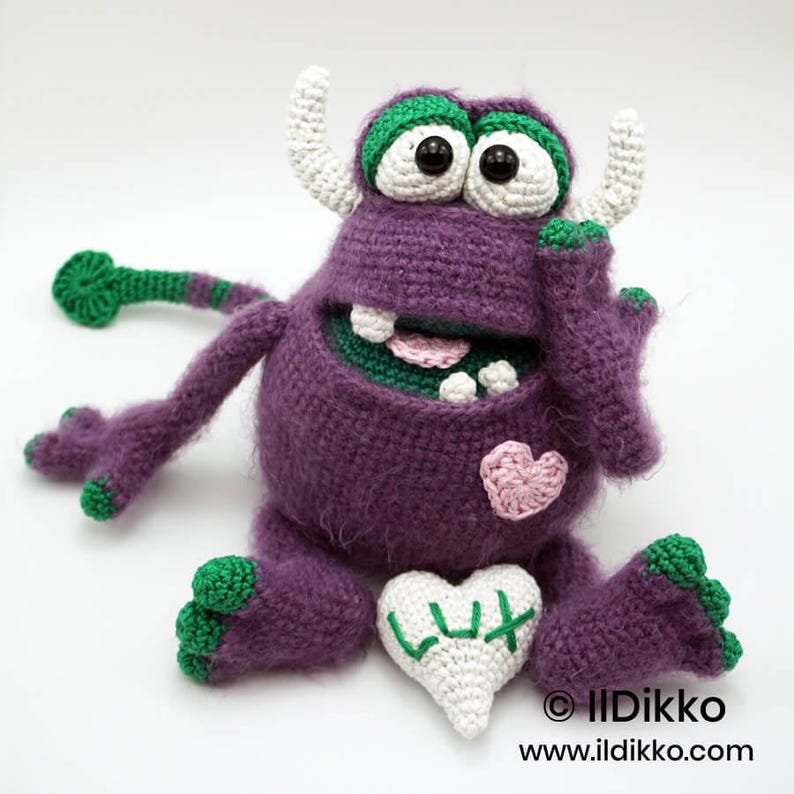 Puede incluir: Un mu&ntilde;eco monstruo de ganchillo morado con detalles verdes, cuernos blancos y ojos grandes. Presenta un coraz&oacute;n rosa en el pecho y sostiene un coraz&oacute;n blanco con el texto bordado en verde LUX. El monstruo tiene una cola larga y extremidades verdes.