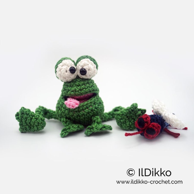 May include: A green crocheted frog with a pink tongue sticking out, looking at a crocheted fly with red eyes and white wings. The frog and fly are on a white background.