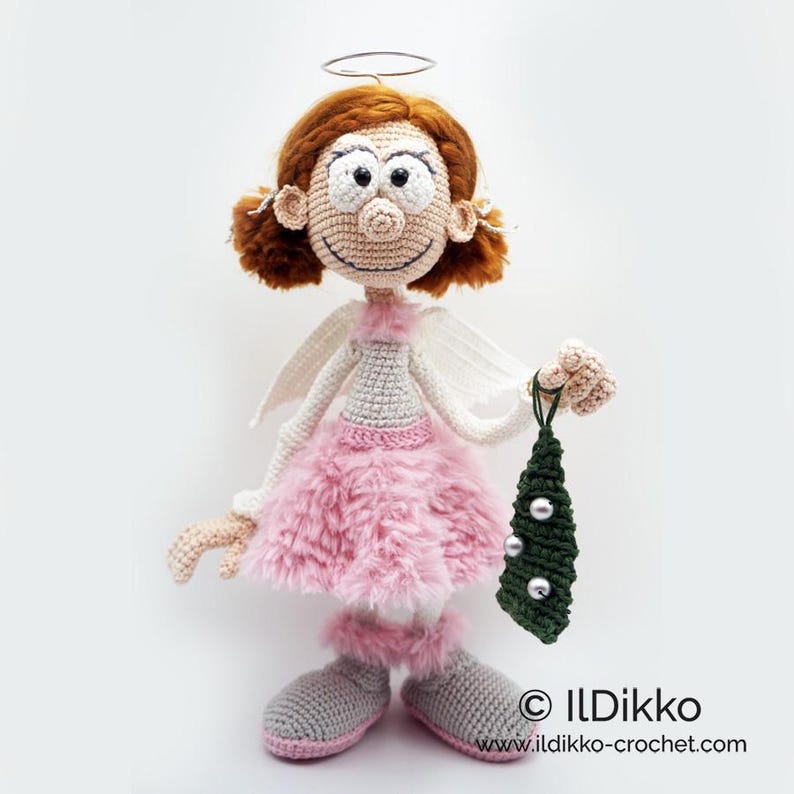 May include: A crocheted doll with auburn hair, a silver halo, and white wings. The doll wears a light pink fuzzy skirt, matching leg warmers, and grey shoes. It holds a small, dark green triangular ornament with three silver beads. Text: ILDikko www.ildikko-crochet.com