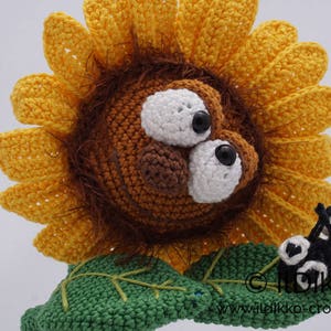 May include: A crocheted sunflower with a brown face and large white eyes. The sunflower has a green stem and leaves. A small black and white crocheted ladybug is sitting on the stem.