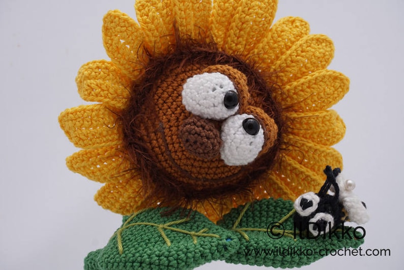 May include: A crocheted sunflower with a brown face and large white eyes. The sunflower has a green stem and leaves. A small black and white crocheted ladybug is sitting on the stem.