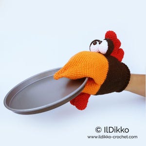May include: A whimsical, handmade oven mitt shaped like a turkey, holding a round, gray baking pan. The mitt features an orange beak, brown head, and red wattle and feathers. The eyes are large and googly. The mitt is holding a round, gray baking pan.
