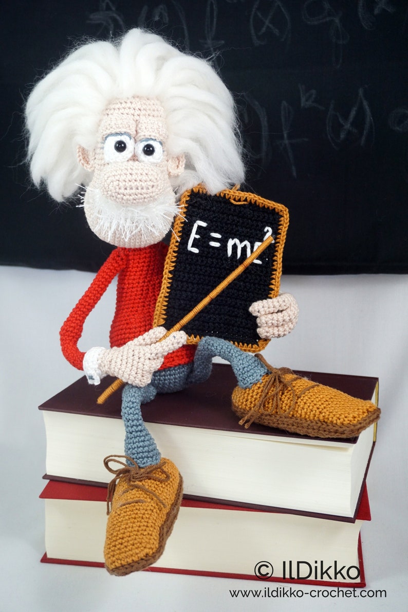Puede incluir: Mu&ntilde;eco de Einstein tejido a ganchillo con pelo y barba blancos, con un su&eacute;ter rojo, pantalones azules y zapatos marrones. Sostiene una peque&ntilde;a pizarra con la ecuaci&oacute;n E=mc&sup2;. El mu&ntilde;eco est&aacute; sentado sobre una pila de libros.