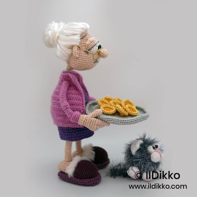 May include: A crocheted doll of a woman wearing a purple sweater, skirt, and slippers, holding a tray of pastries. A gray cat with white accents sits nearby. The doll has white hair and glasses. The tray has a gray border and the pastries are yellow.