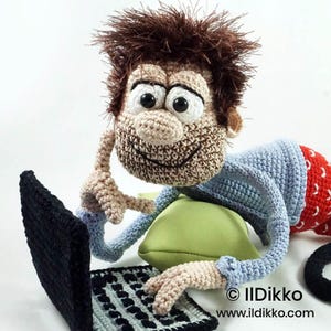 May include: A crocheted doll with spiky brown hair, large white eyes, and a tan face, wearing a light blue long-sleeved shirt and red patterned pants. The doll is lying on a light green pillow, using a black and gray crocheted laptop computer.