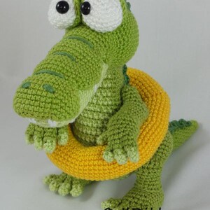 May include: A green crocheted crocodile toy with large white eyes and black pupils, wearing a bright yellow crocheted swim ring around its waist. The text "&copy; ILDikko www.ildikko-crochet.com" is printed in black at the bottom of the image.