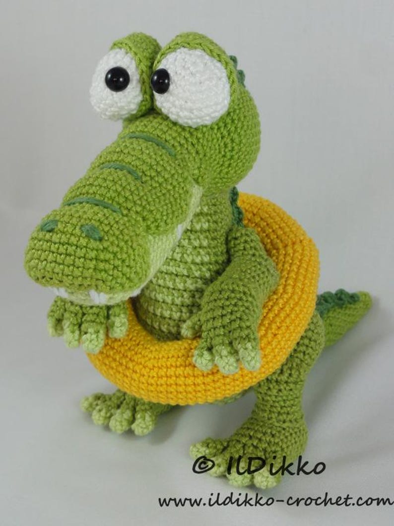 May include: A green crocheted crocodile toy with large white eyes and black pupils, wearing a bright yellow crocheted swim ring around its waist. The text "&copy; ILDikko www.ildikko-crochet.com" is printed in black at the bottom of the image.