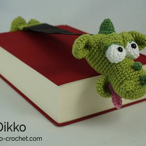 May include: A green crocheted dragon bookmark with white eyes and a pink tongue, peeking out from a red book.