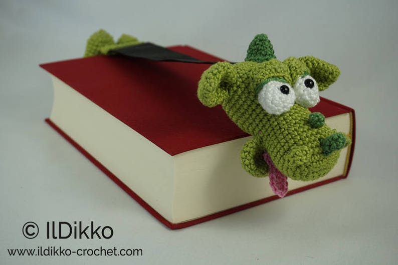 May include: A green crocheted dragon bookmark with white eyes and a pink tongue, peeking out from a red book.