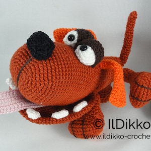 May include: A crocheted orange dog toy with a pink tongue sticking out. The dog has black eyes and a brown nose. The toy is sitting on a white surface.