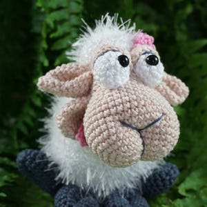 May include: A crocheted sheep toy with white fur, pink ears, and blue feet. The sheep has large, white eyes with black pupils and a pink nose.