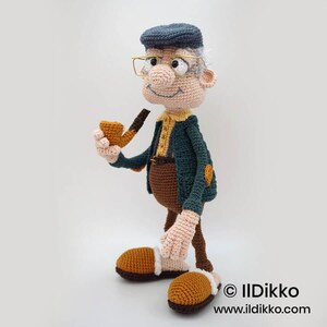 May include: A handmade crochet doll of an elderly man wearing a blue cap, glasses, and a green jacket. The doll is holding a brown pipe and has brown pants and shoes. The doll has a smiling expression and is made with various yarn colors.
