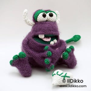 Puede incluir: Un monstruo de ganchillo morado con dos cuernos blancos, ojos grandes blancos con pupilas negras y detalles verdes en pies y manos. El monstruo tiene una boca ancha con dientes visibles y una lengua rosa, sentado junto a un objeto blanco en forma de coraz&oacute;n.