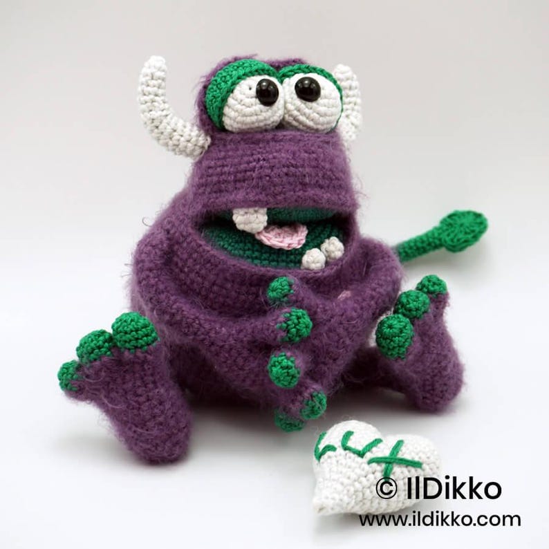 Puede incluir: Un monstruo de ganchillo morado con dos cuernos blancos, ojos grandes blancos con pupilas negras y detalles verdes en pies y manos. El monstruo tiene una boca ancha con dientes visibles y una lengua rosa, sentado junto a un objeto blanco en forma de coraz&oacute;n.