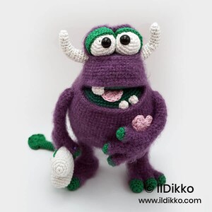 Puede incluir: Un monstruo de ganchillo morado con dos cuernos blancos, ojos grandes blancos con pupilas negras y p&aacute;rpados verdes. Tiene una boca ancha con dientes blancos y una lengua rosa, sosteniendo un peque&ntilde;o coraz&oacute;n rosa. Texto Ildikko ildikko.com.