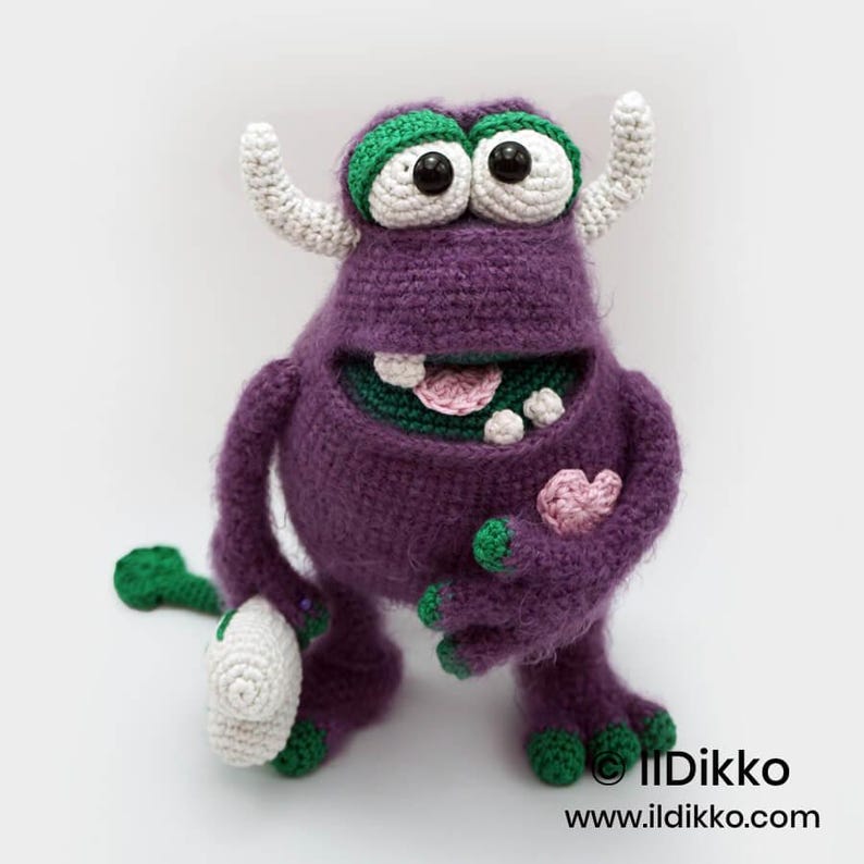 Puede incluir: Un monstruo de ganchillo morado con dos cuernos blancos, ojos grandes blancos con pupilas negras y p&aacute;rpados verdes. Tiene una boca ancha con dientes blancos y una lengua rosa, sosteniendo un peque&ntilde;o coraz&oacute;n rosa. Texto Ildikko ildikko.com.