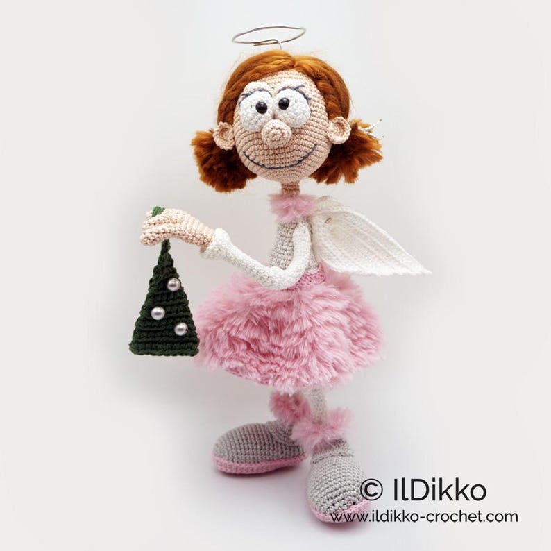 May include: A crocheted doll with auburn hair, a wire halo, and white wings, wearing a light gray top and a fluffy pink skirt. The doll has gray shoes with pink trim and holds a small dark green triangular ornament with white beads. Text: ILDikko.