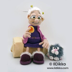 May include: Crocheted doll of a smiling elderly woman wearing glasses, a purple dress, and a pink jacket, holding a yellow cookie. A small gray cat doll sits beside her. The doll has white hair and is wearing purple slippers.
