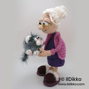 May include: Crocheted doll of a woman holding a gray cat. The woman wears a purple dress, pink sweater, and brown slippers. The cat is gray with pink accents. The doll has white hair and glasses. The doll is handmade.