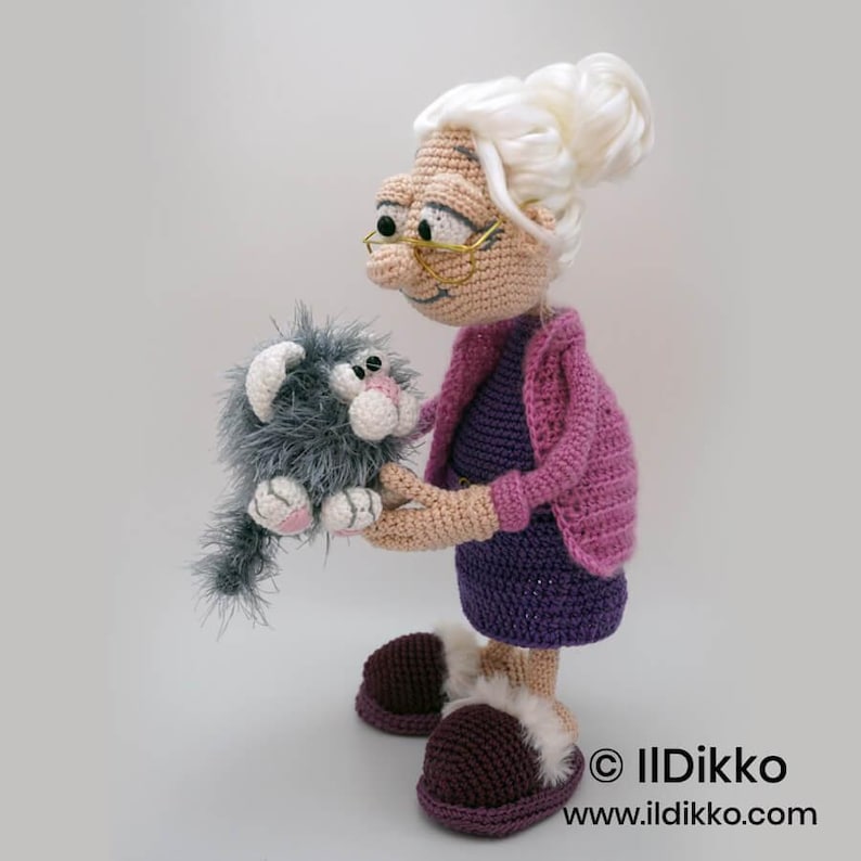 May include: Crocheted doll of a woman holding a gray cat. The woman wears a purple dress, pink sweater, and brown slippers. The cat is gray with pink accents. The doll has white hair and glasses. The doll is handmade.