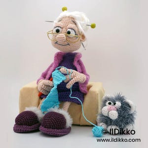 May include: A handmade crochet doll of a grandmother wearing glasses, a purple dress, and a pink jacket, knitting a blue scarf. A gray cat doll sits nearby with a ball of blue yarn. The doll is wearing purple slippers with white trim.