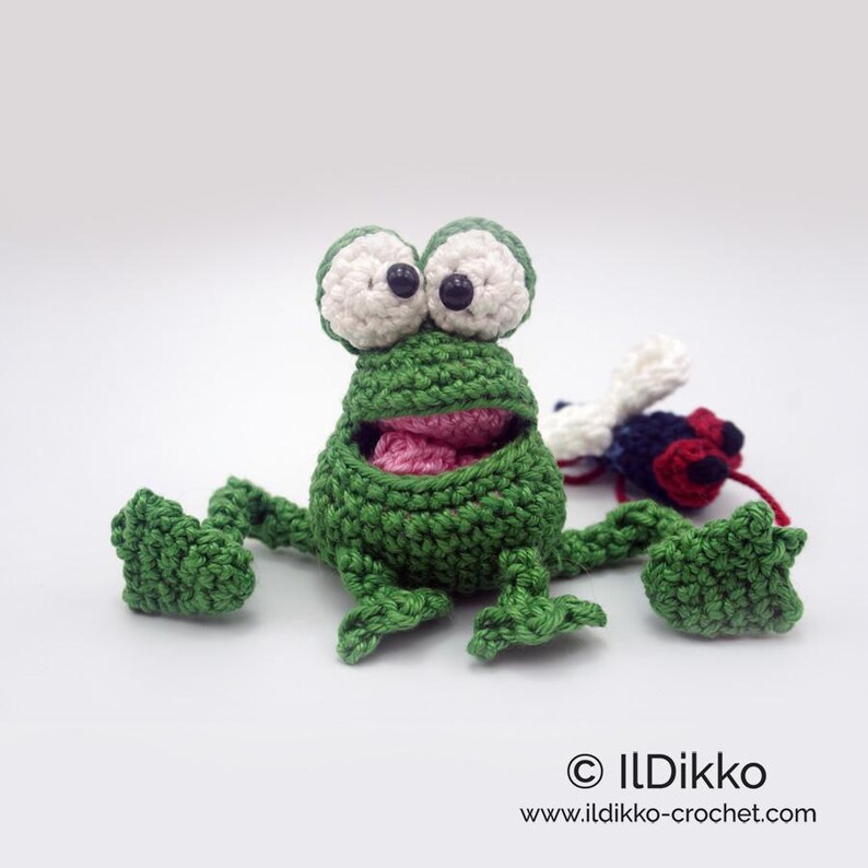 May include: A green crocheted frog with large white eyes and a pink mouth, holding a crocheted fly in its mouth. The frog is sitting on a white surface.