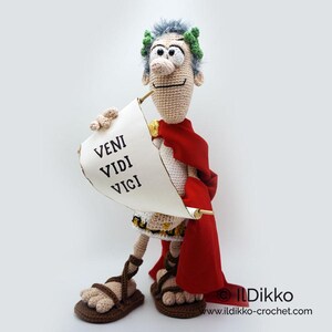 以下が含まれることがあります： 「VENI VIDI VICI」と書かれた巻物を手に持つ、ローマ皇帝の編みぐるみ人形。赤いトガと月桂樹の冠を身に着けている。