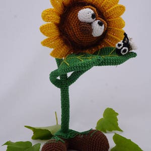 May include: A crocheted sunflower with a brown lion face and large eyes. The sunflower has a green stem and leaves. A small black and white crocheted bug is perched on a leaf. The sunflower is standing on a white surface with green leaves.
