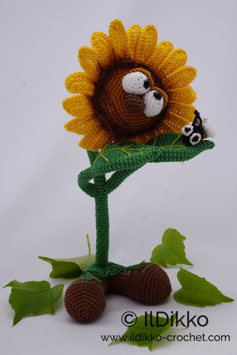 May include: A crocheted sunflower with a brown lion face and large eyes. The sunflower has a green stem and leaves. A small black and white crocheted bug is perched on a leaf. The sunflower is standing on a white surface with green leaves.