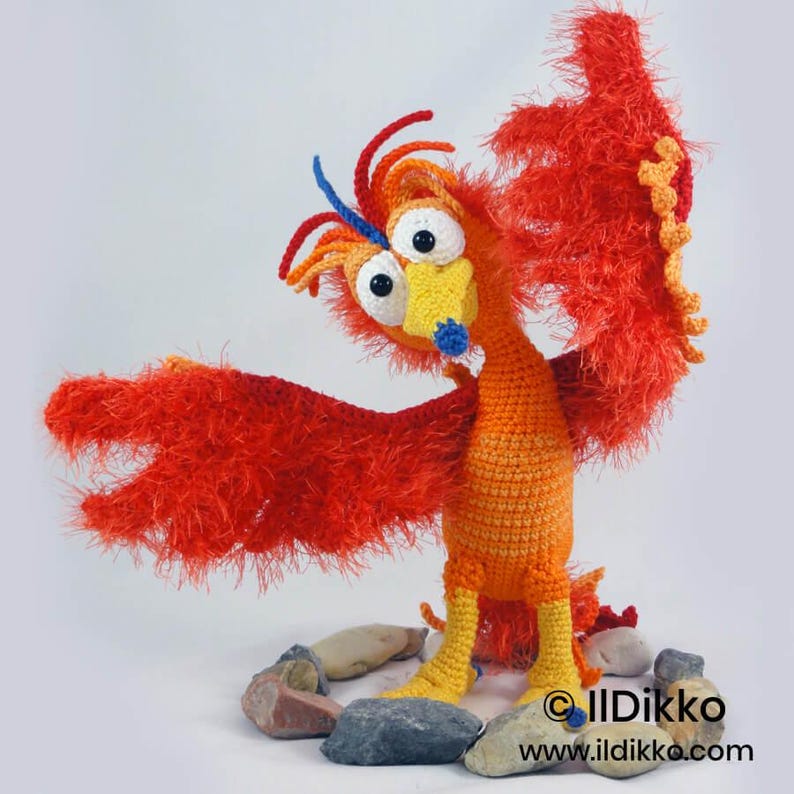 May include: A crocheted phoenix figure with bright orange and red fuzzy yarn wings, a yellow beak with a blue tip, and large white eyes with black pupils. The creature stands on a base of smooth gray stones. Text reads: IlDikko www.ildikko.com.
