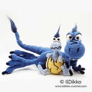 May include: A blue crocheted dragon toy with a long tail and black claws, accompanied by a smaller light blue baby dragon emerging from a yellow cracked egg. The text "&copy; ILDikko www.ildikko-crochet.com" is printed in the bottom right corner.