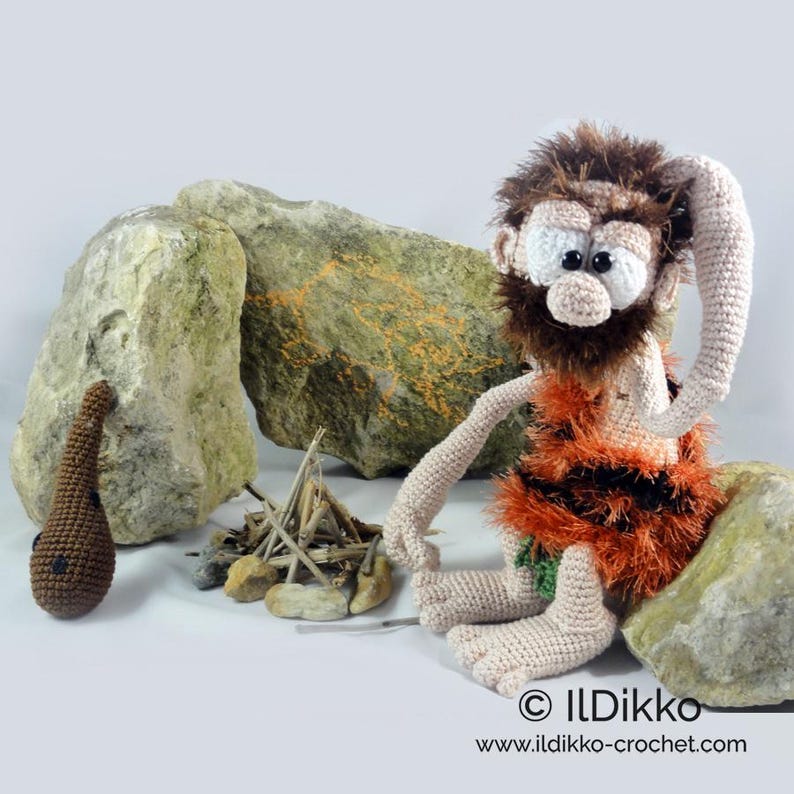 May include: A crocheted caveman doll with brown hair and beard, wearing an orange and black fuzzy tunic. The doll sits beside a small pile of sticks and a brown crocheted club. Text at the bottom reads ILDikko and www.ildikko-crochet.com.