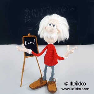May include: A crocheted doll of a scientist with wild white hair, a red long-sleeved shirt, and gray pants. The doll stands next to a small wooden easel holding a black chalkboard with the white text E=mc2 written on it. The doll wears brown shoes.