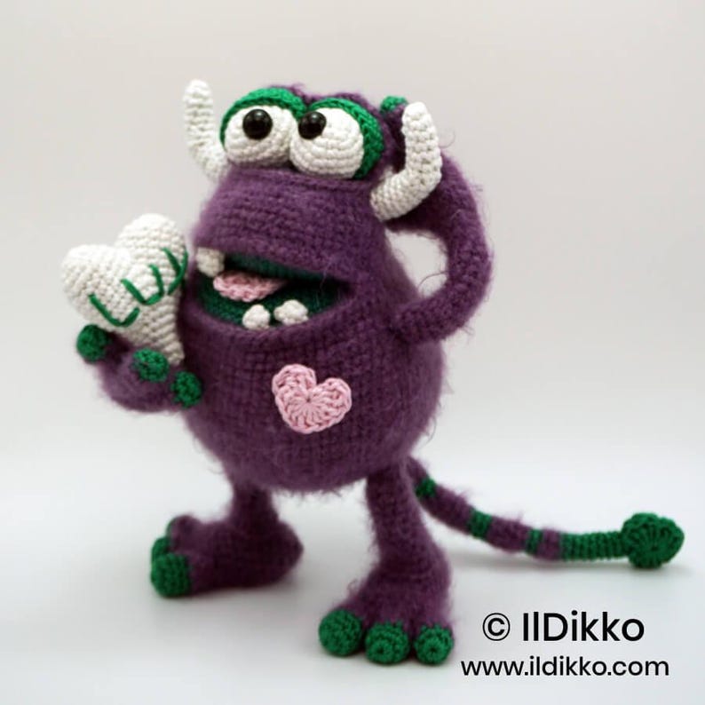 Puede incluir: Un monstruo de ganchillo morado con detalles verdes, cuernos blancos y ojos grandes. Sostiene un coraz&oacute;n blanco con el texto LUV y tiene un peque&ntilde;o coraz&oacute;n rosa en el pecho. La criatura tiene una cola larga a rayas y pies y manos verdes.
